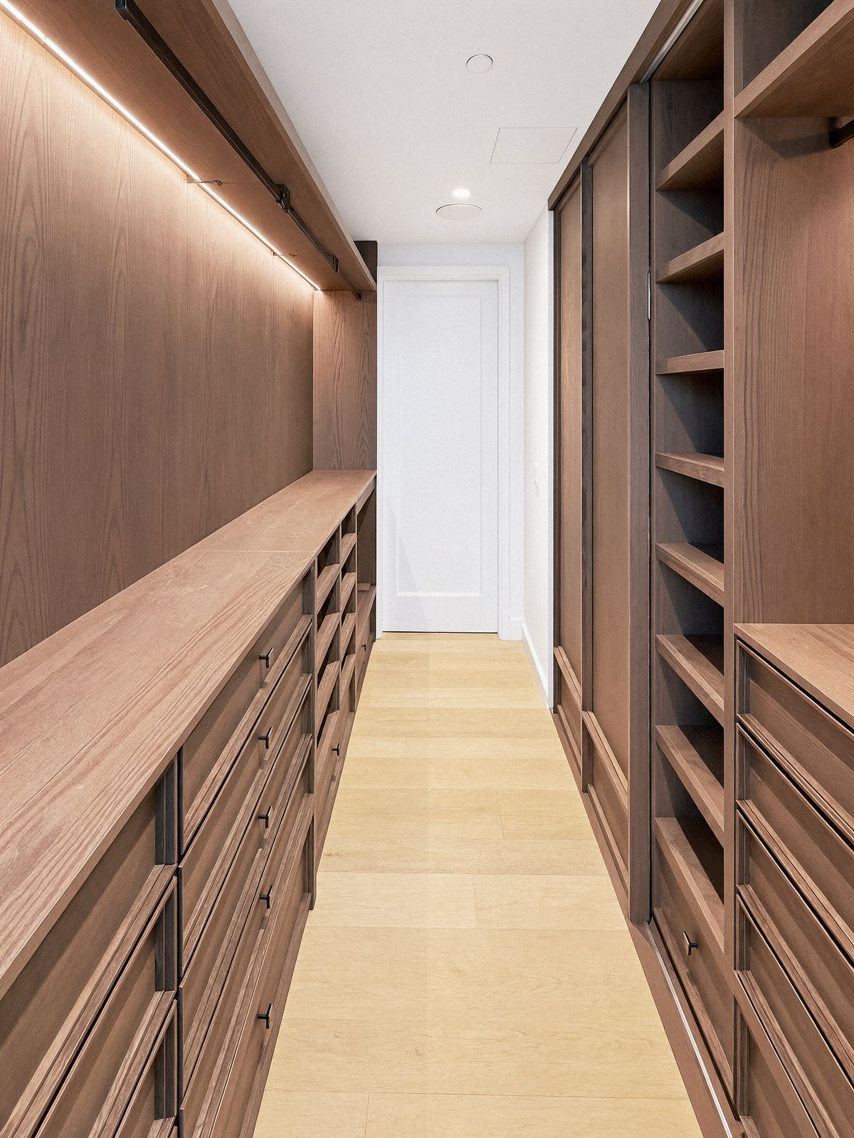 walk-in closet corridor with panelled oak wardrobes on one side, a counter over drawers on the other and a door at the end